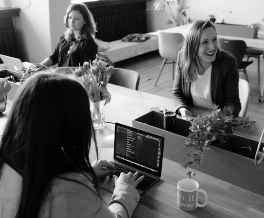 Group of 3 women inside an office talking. They have laptops